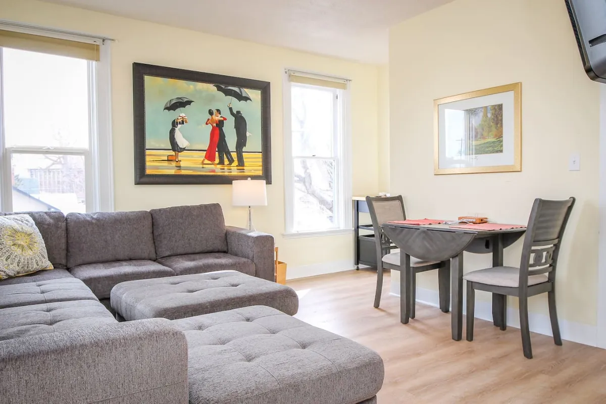The image shows a cozy, art-filled living and dining area inside a beautifully restored Victorian apartment. A plush sectional sofa invites relaxation, while a stylish dining table and chairs offer space for meals or conversation. Eye-catching artwork on the wall adds a creative, historic flair, setting the scene for memorable gatherings.