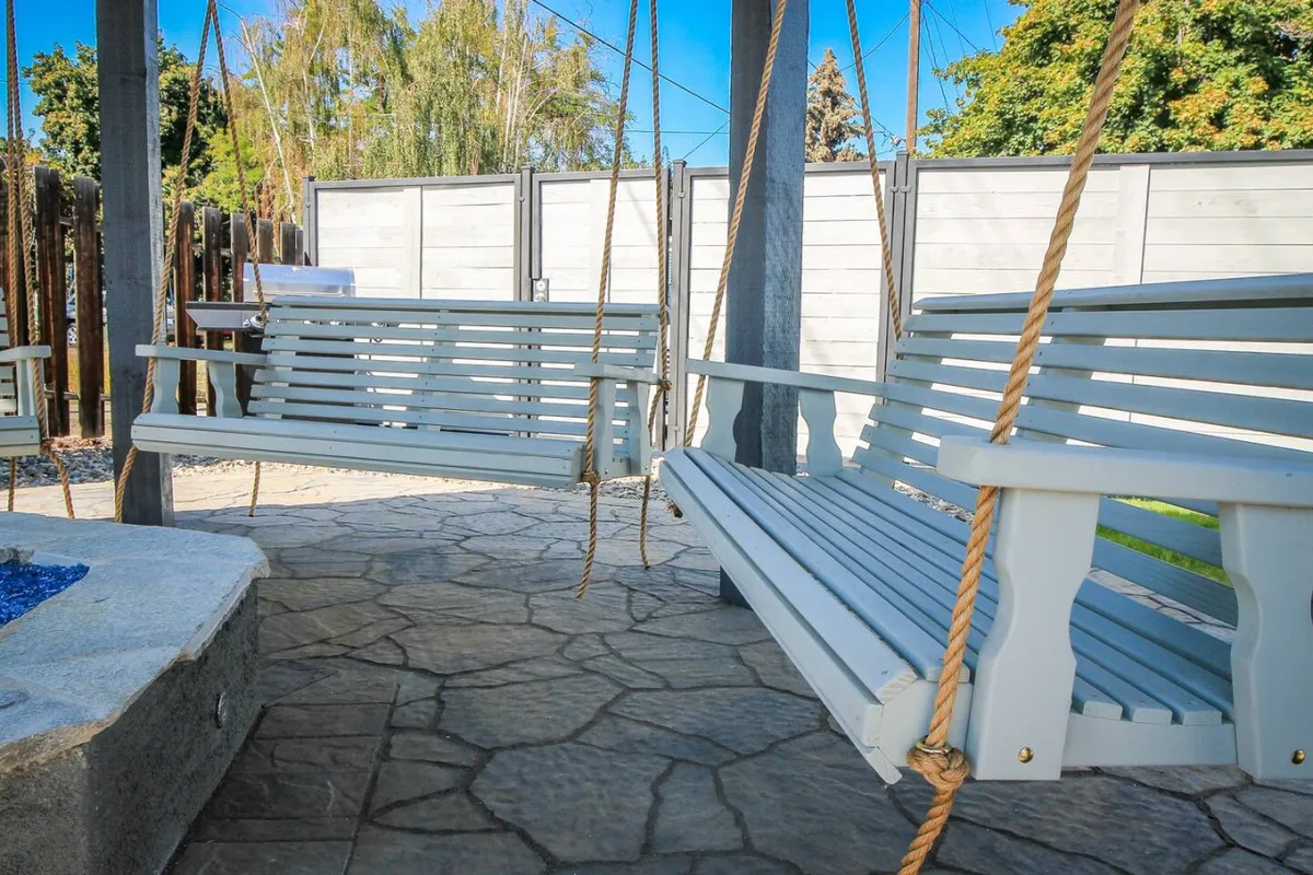 This image captures a communal backyard scene featuring inviting swing seats, a cozy fire pit, stone flooring, and lush greenery surrounding the area. The ambiance is tranquil and welcoming, with spaces for socializing and relaxation beneath the trees.