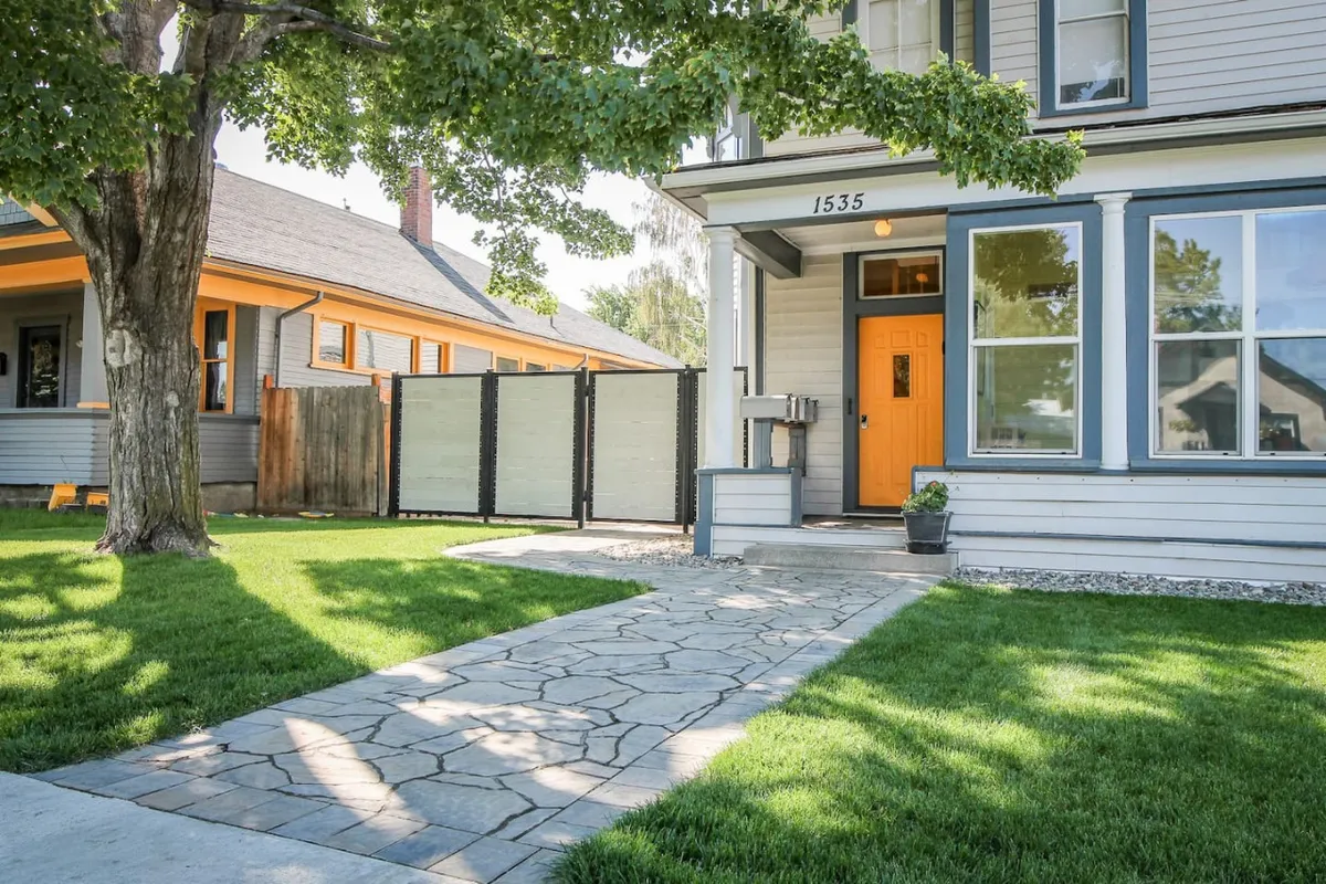 The exterior of Palmer House features a bright orange door, a stone pathway bordered by a lush green lawn, and a large shady tree. The inviting front yard hints at a welcoming return after a day outdoors.