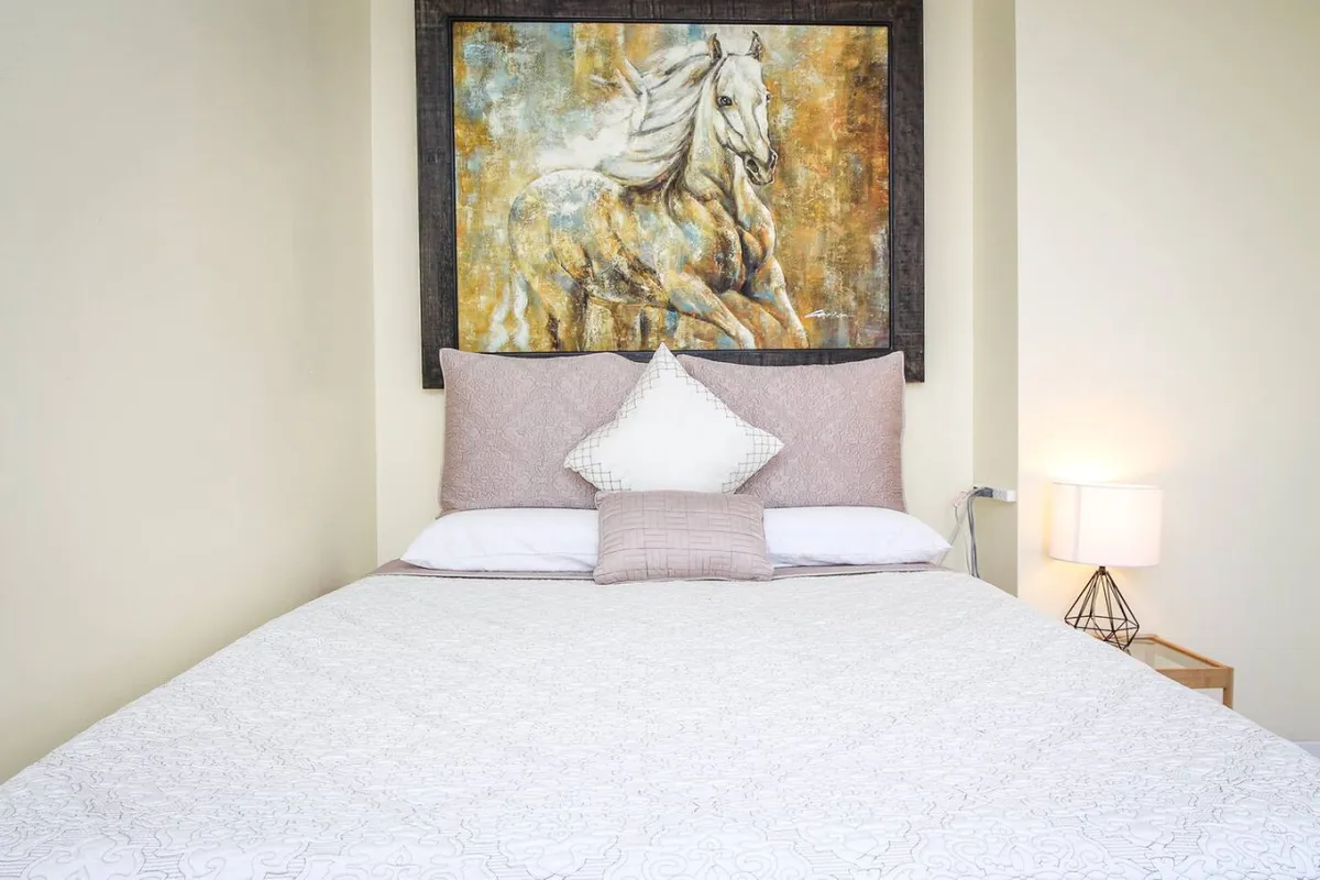 This image presents a tranquil sleeping nook with a neatly made bed featuring soft linens and decorative pillows. A horse painting above the bed adds a touch of artistry, while a table lamp on the nightstand suggests a cozy environment for reading or relaxing before sleep. Morning light filters into the room, enhancing its peaceful ambiance.
