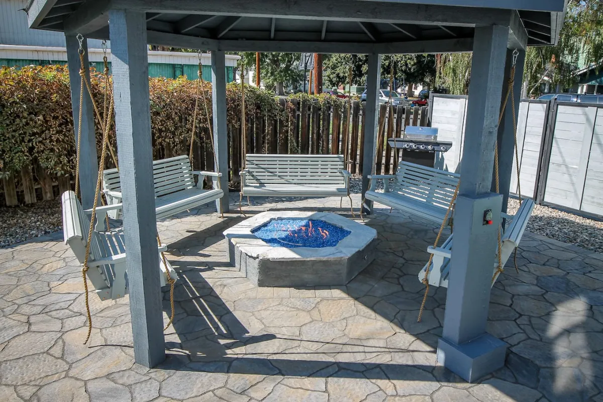 This image showcases a communal backyard setting featuring a cozy fire pit, hanging swings, and a stone patio. The scene is set for guests to relax together, gather around the fire, and enjoy the outdoors in a tranquil atmosphere.