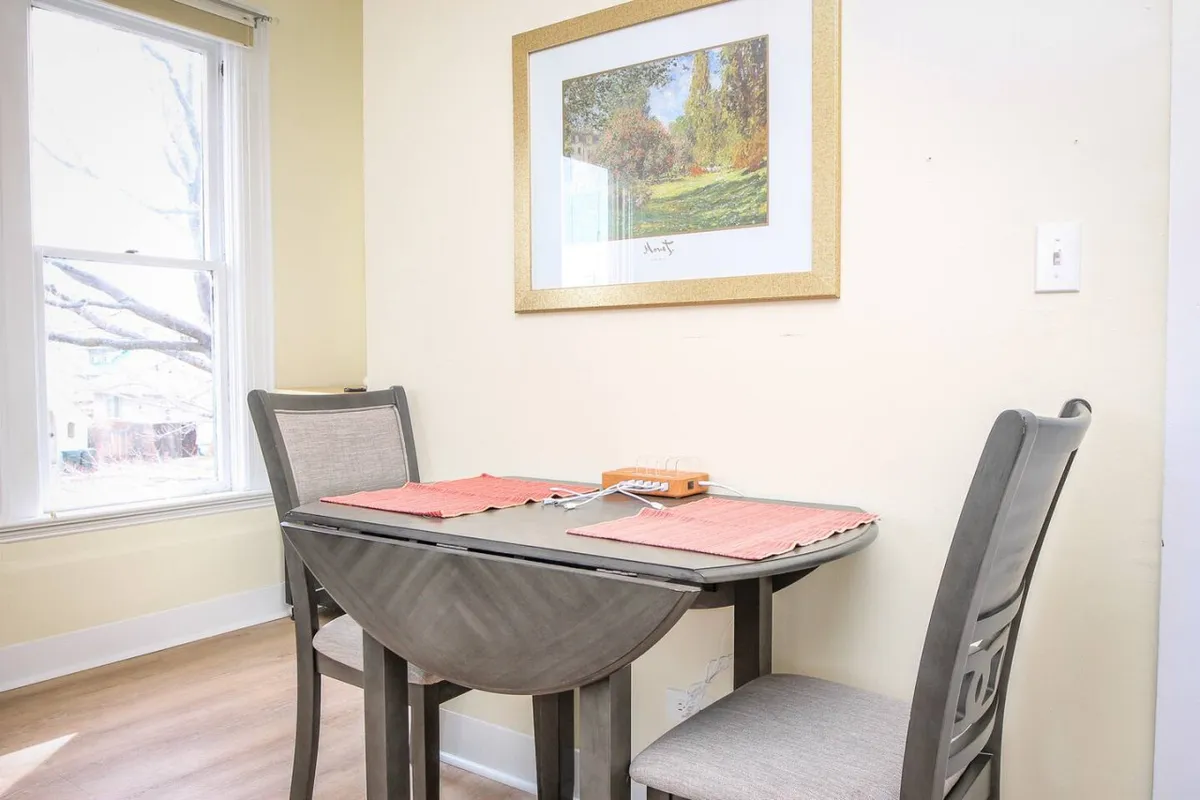 This image features a small, cozy dining nook with a table for two set beside a window awash in natural light. Framed artwork hangs on the wall, and the space radiates warmth and intimacy, ideal for quiet meals or heartfelt conversation.