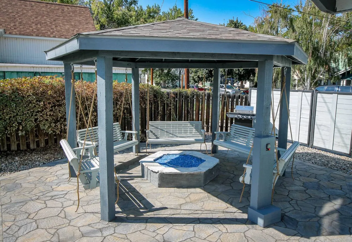 A gazebo structure anchors this backyard scene, surrounded by swings and a fire pit adorned with blue stones. The space is designed for socializing, relaxing, and sharing meals under the open sky, highlighting a comfortable and communal atmosphere.